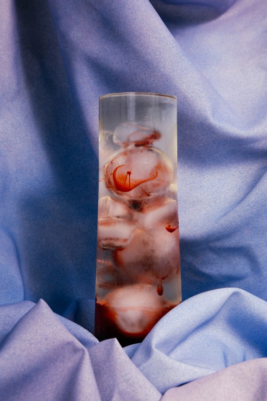 Against a lavender fabric background stands a cylinder filled with a clear liquid and bulbous shapes that appear to be bloody and tissue specimen-like.