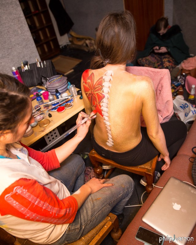 Image of the back of a topless person with long dark hair, having a spine and flowers painted on their back. The painter is wearing a red and white top, jeans and is holding a paint brush with red paint. There are painting supplies on the table next to the two people and a person in the background looking at their phone.