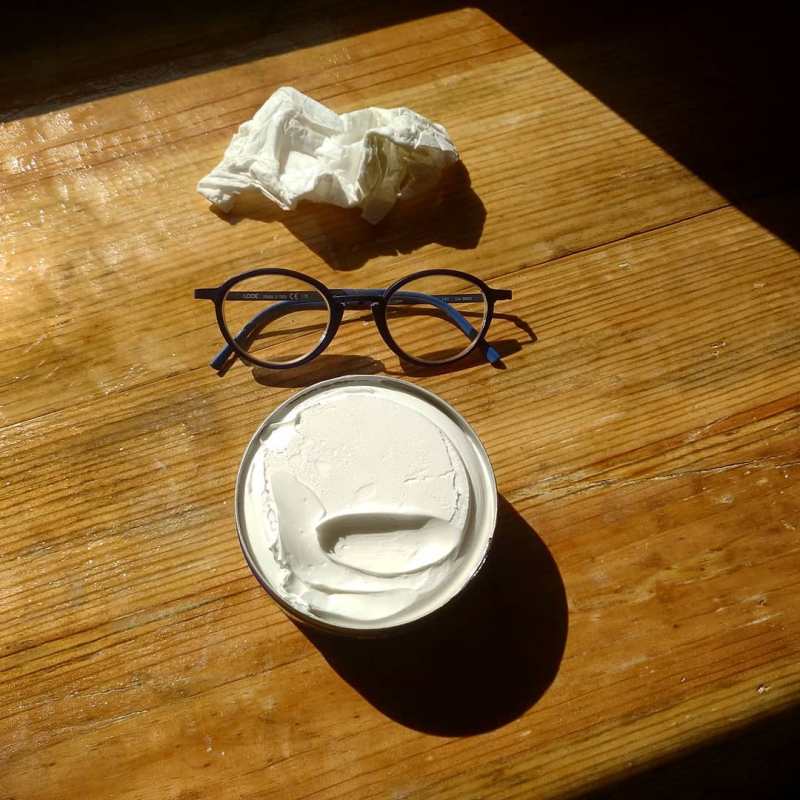 Photo of a crumped up tissue, a pair of glasses and a jar of white cream on a wooden table