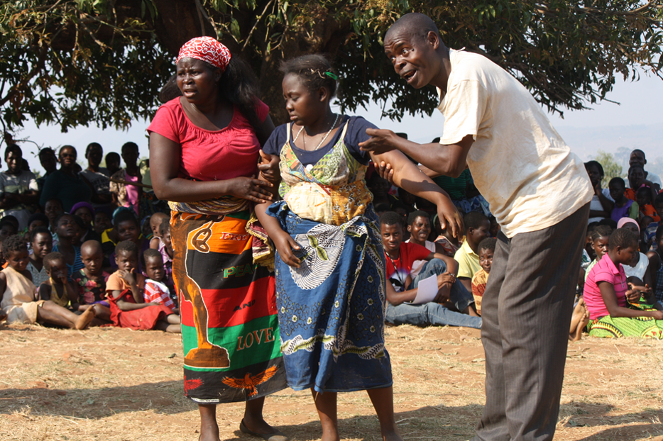 A man and a woman hold up a second woman by her arms, whilst a crowd of people seated in the background looks on