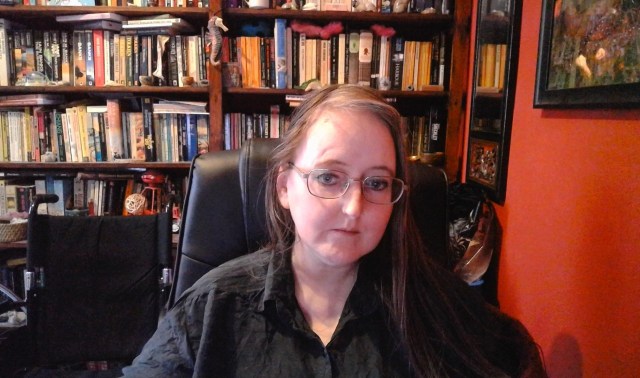 Image of a person with long, light-coloured hair sat on a black desk chair, wearing glasses and a black top. The person is sat next to an orange wall, with a framed picture and mirror. The background is a set of bookshelves full of books, and other items. A wheelchair with a black seat and handles is visible to the left of the image.