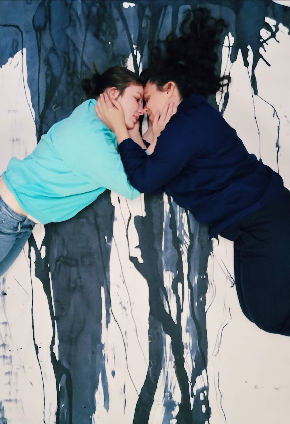 Image of two people, lying on the floor against a blue and white background. The people are lying with their heads together, holding each others head with their hands. Their bodies are directed away from each other.