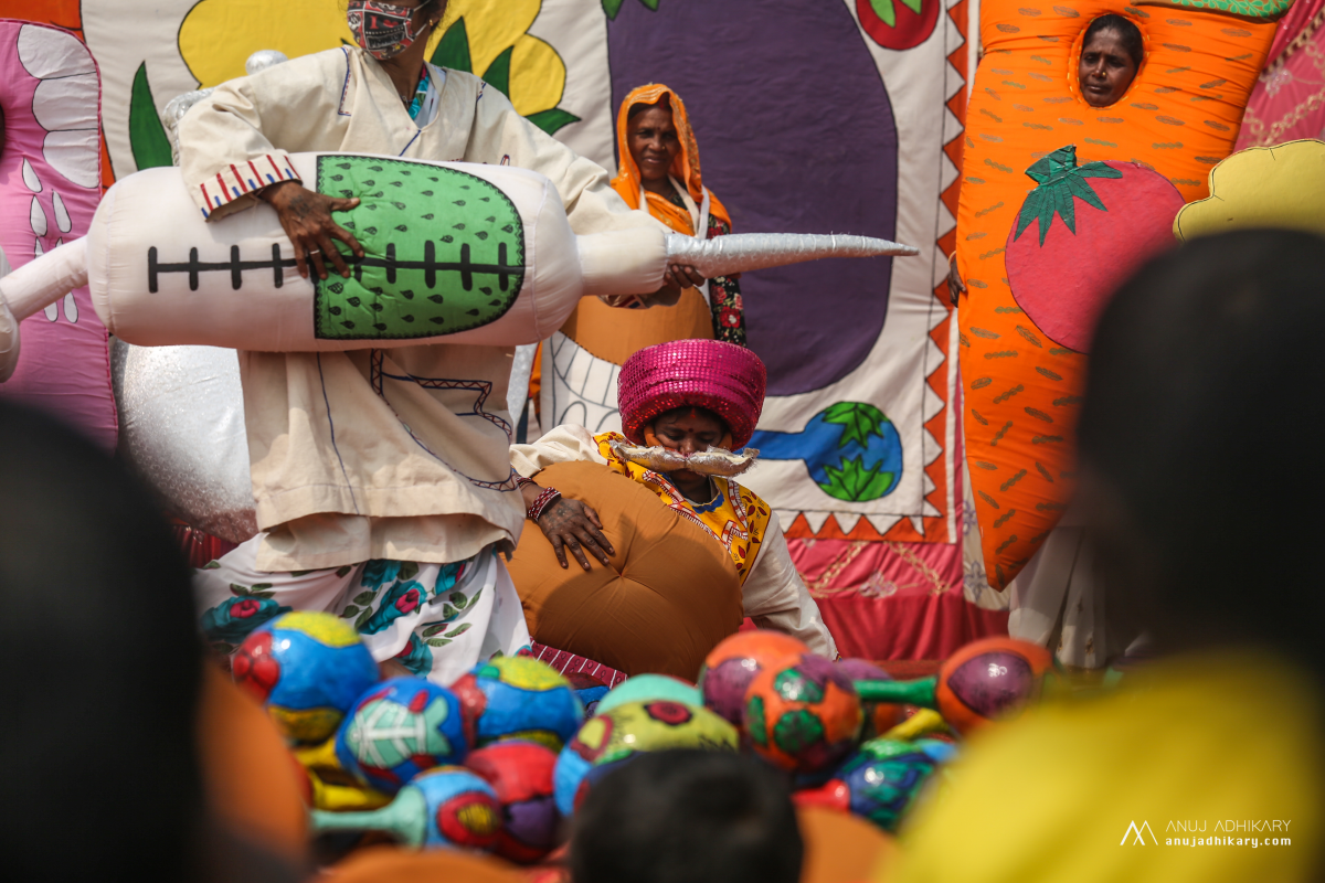 Colourful image of people dressed in costumes, holding a large needle. One person is fallen on the floor wearing a large belly costume.