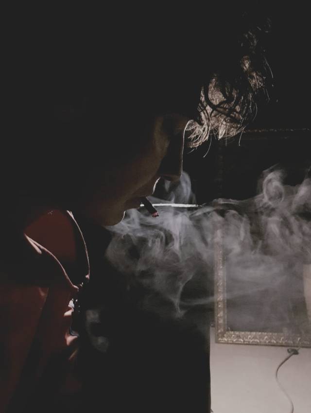 Image of a person with curly hair with a cigarette in their mouth, surrounded by smoke. The lighting is from behind and lights up some of the hair and the edges of the persons cloth