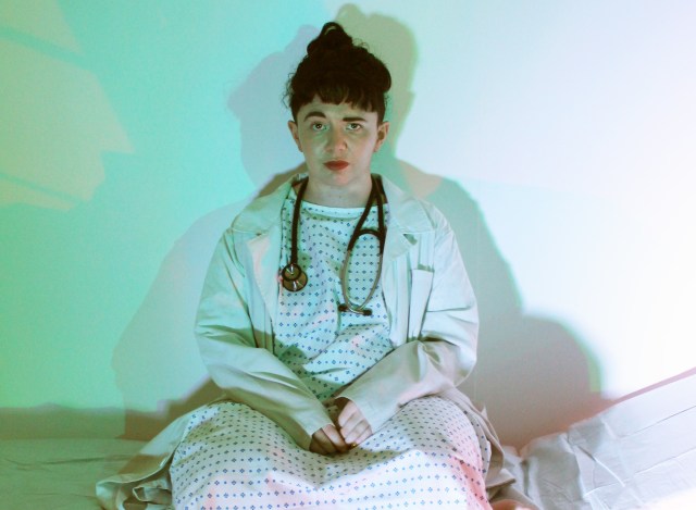 A person with dark hair and red lipstick sits the edge of an examination table, wearing a hospital gown, a white doctor's coat and a stethoscope around their neck