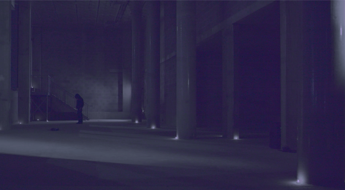 A dark photograph, of a person standing at the back of a room. The space is made of concrete with tall pillars to the side. There are small candles or lights at the base of each of these pillars casting a soft purple glow. There is a shaft of light that falls across the floor and cuts diagonally across the image. The figure at the back appears all in black, standing, with their head slightly bent forward.